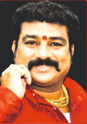 Smiling Indian man with a mustache wearing a red shirt and gold jewelry resting his chin on his hand